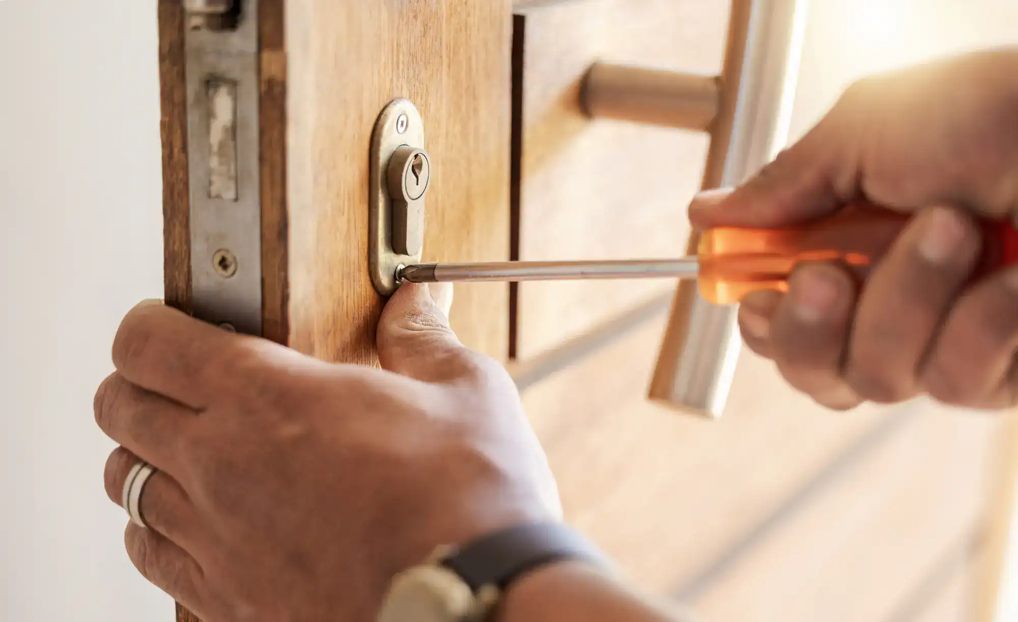 A person uses a screwdriver to install or repair a lock on a wooden door, tightening a screw near the lock mechanism—showcasing skilled locksmith services NYC. Only the person's hands and part of the door handle are visible.