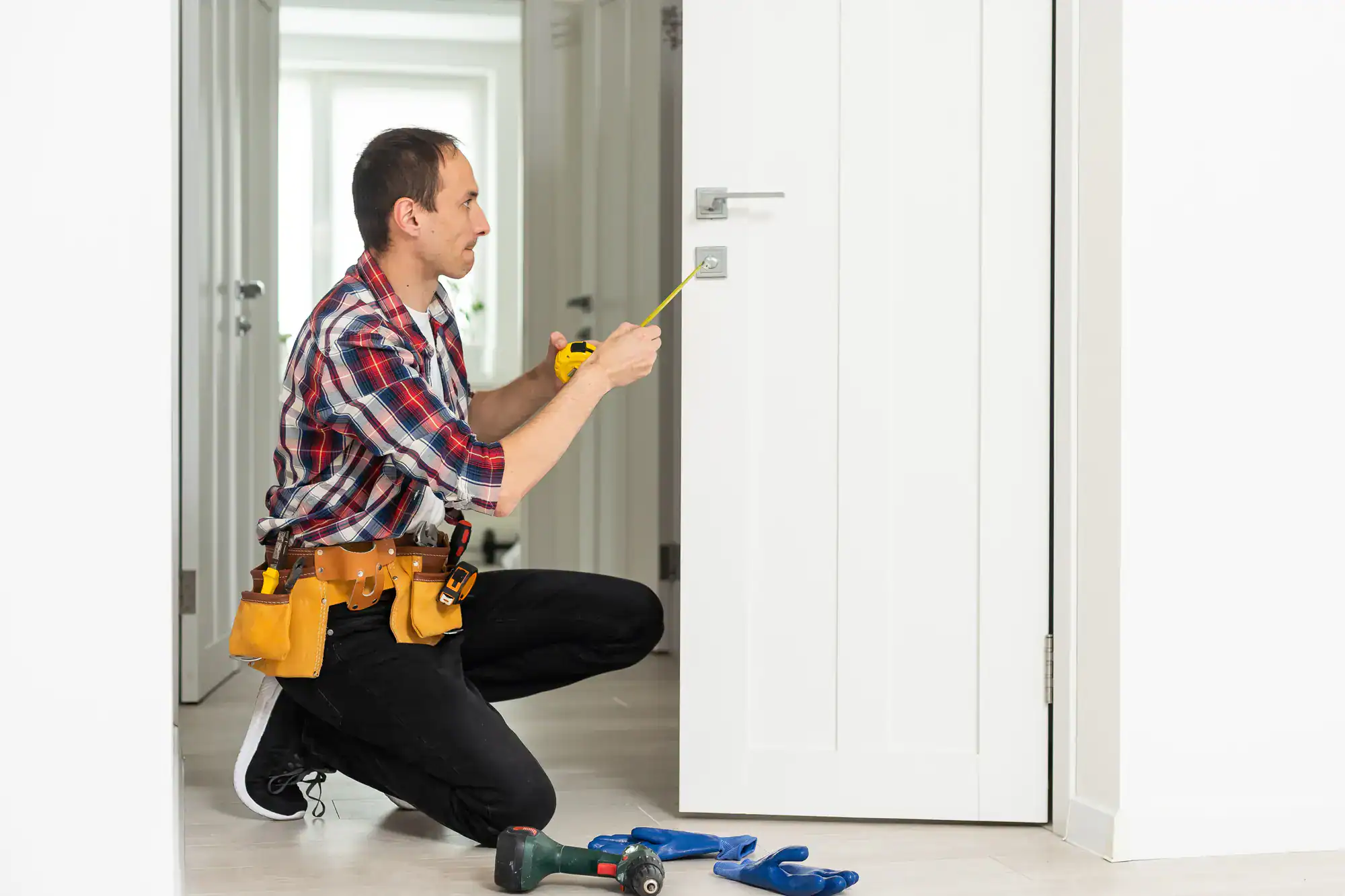 A man wearing a tool belt kneels beside a white door, using a tape measure to check the handle area. Various tools are laid out on the floor nearby, highlighting the precision of locksmith services NYC in a bright, modern interior.