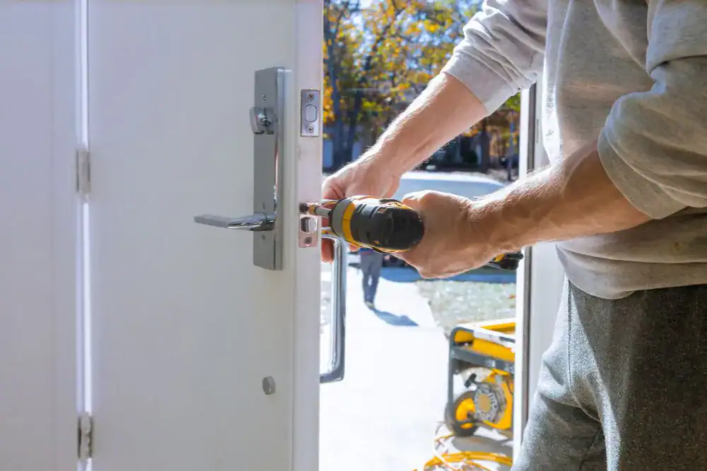 A person uses a power drill to install or repair a lock on a white door, with sunlight and outdoor scenery visible in the background—showcasing expert locksmith services NYC.