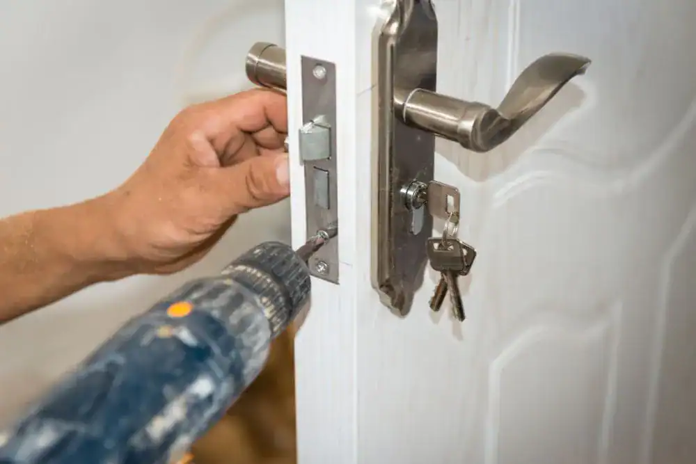 A person uses a power drill to install or repair a metal door lock on a white door, with a set of keys hanging from the lock—showcasing skilled locksmith services NYC residents can trust.