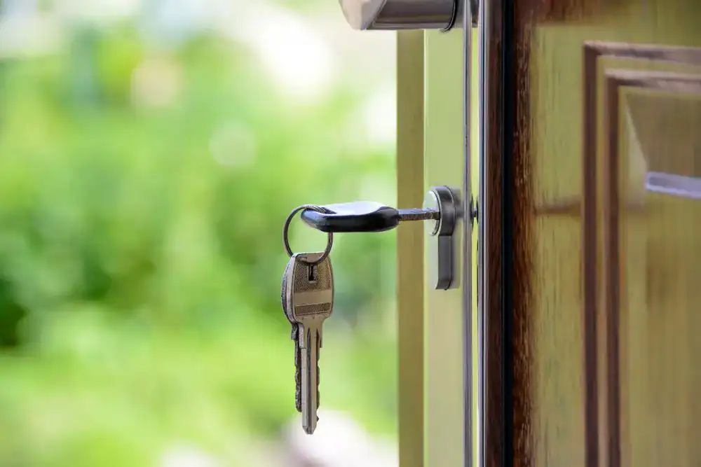 A close-up of a key inserted into a door lock, with two keys on the keyring. The wooden door is slightly open, greenery blurred outside—a scene often seen in professional locksmith services NYC.