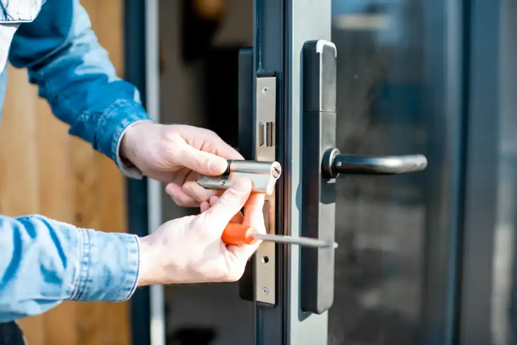 A person in a blue shirt uses tools to install or repair a door lock cylinder on a modern glass door, showcasing skilled locksmith services NYC with one hand holding the lock and the other using a screwdriver.