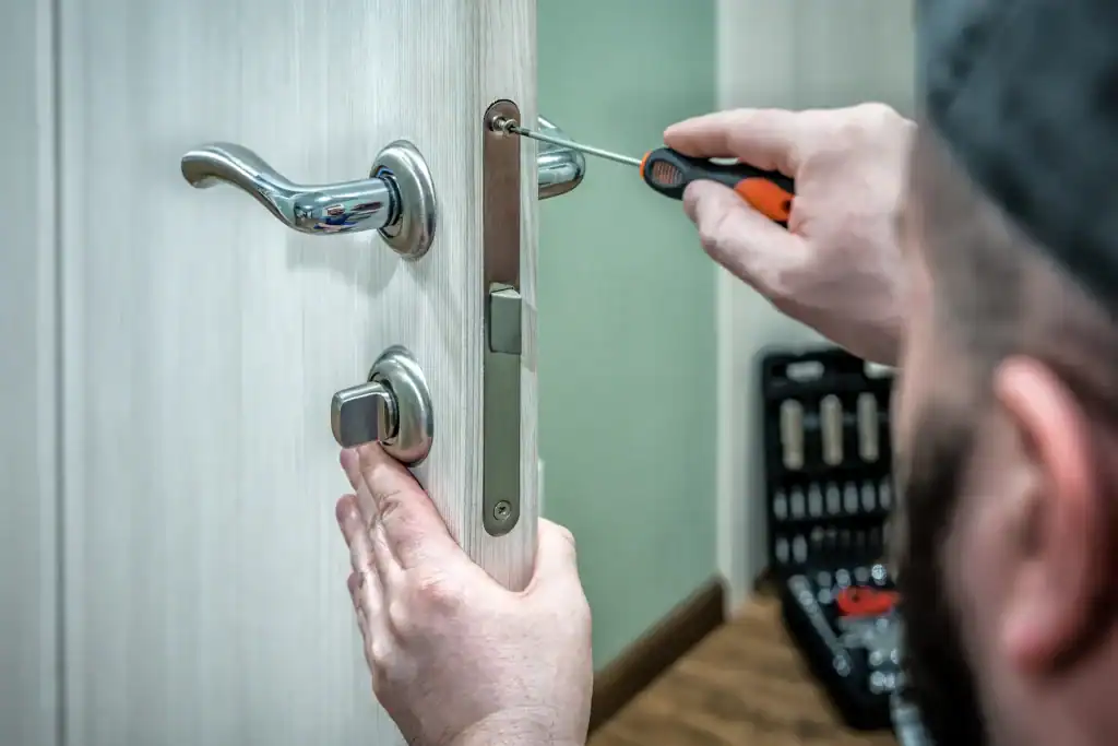 A person using a screwdriver to fix or install a door lock on a light-colored wooden door, with a toolkit visible in the background, showcasing the expertise of locksmith services NYC.