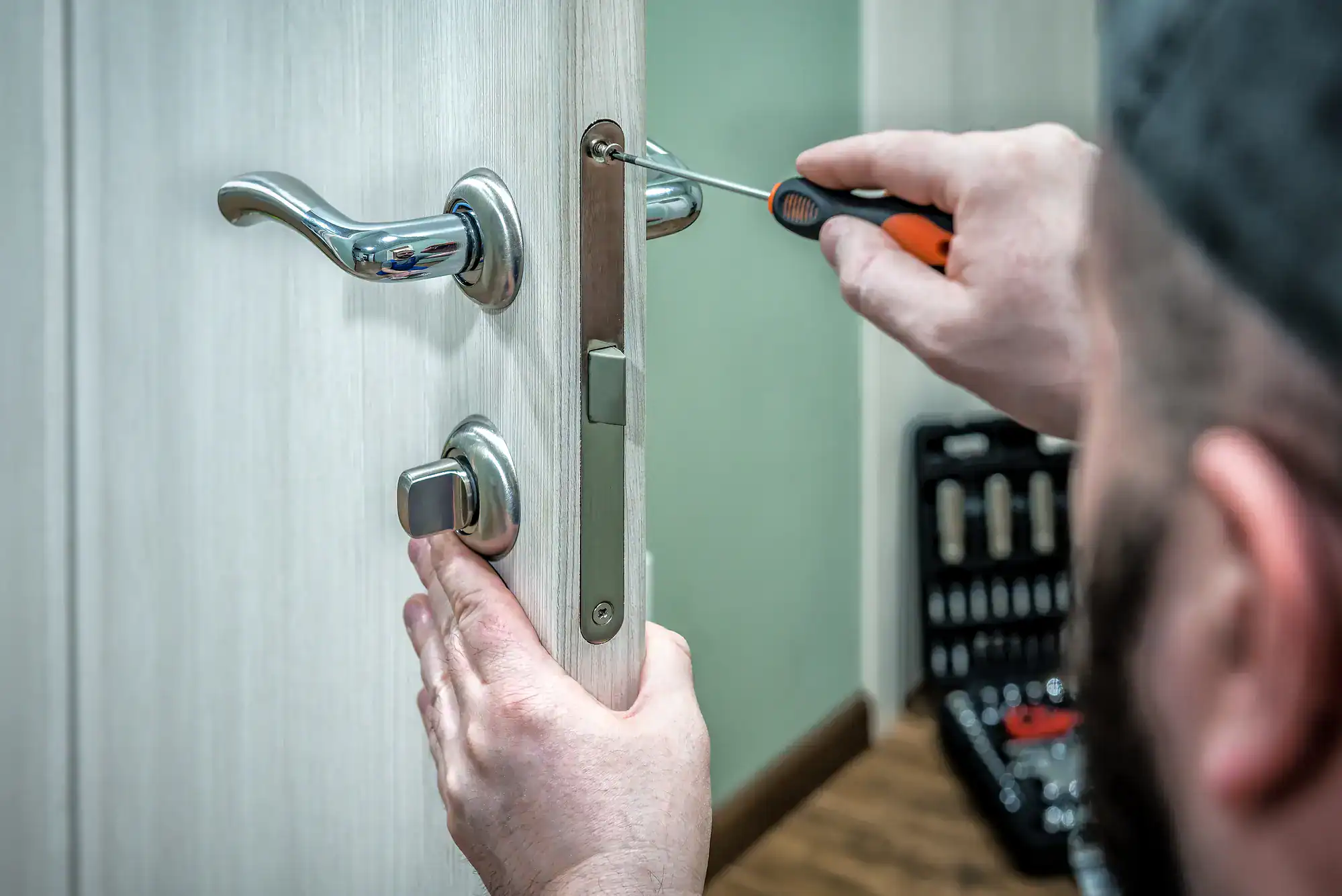 A person using a screwdriver to fix or install a door lock on a light-colored wooden door, with a toolkit visible in the background, showcasing the expertise of locksmith services NYC.