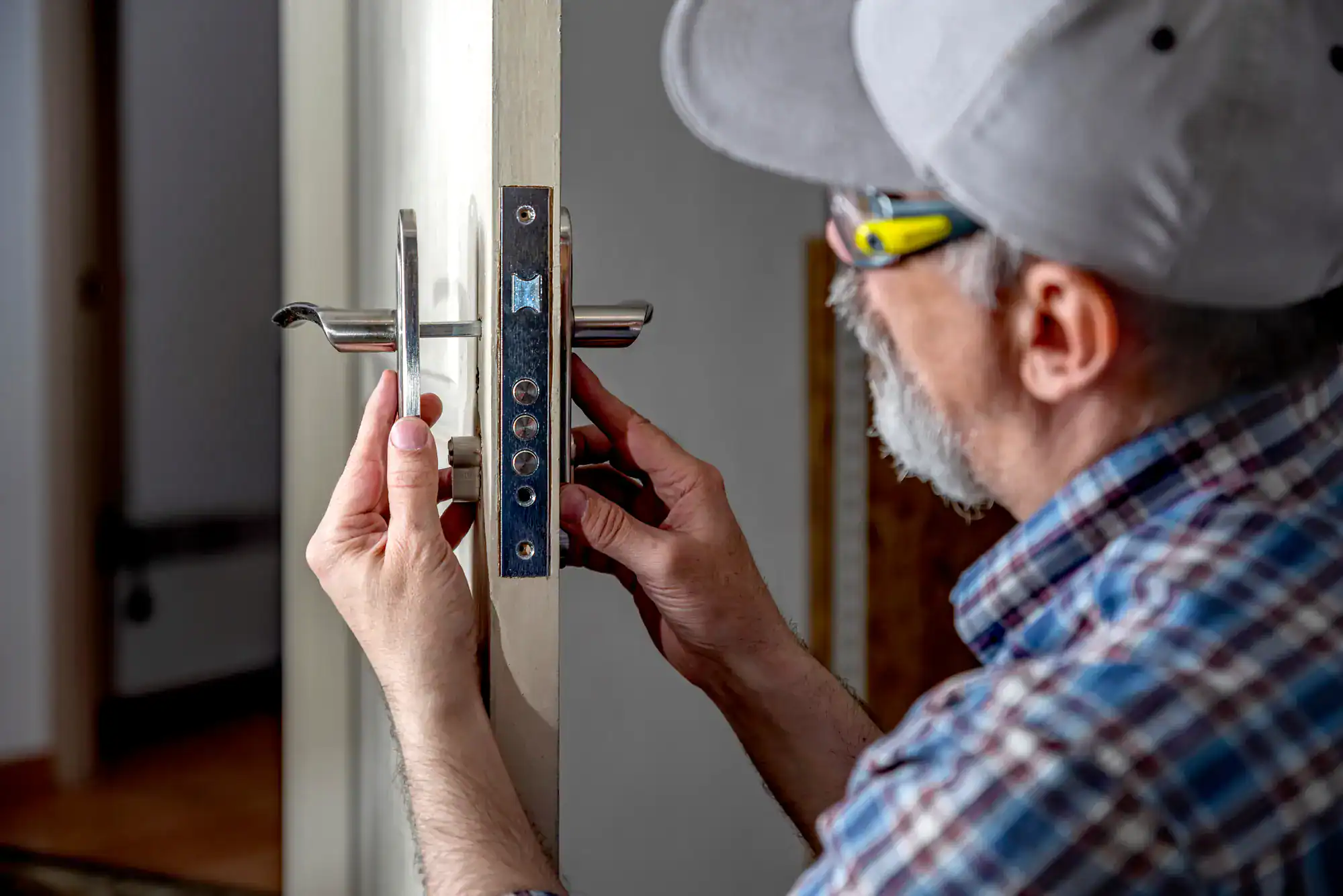 A man wearing a cap and glasses installs or repairs a door handle and lock on a white door, using a screwdriver—demonstrating professional locksmith services NYC residents can rely on.