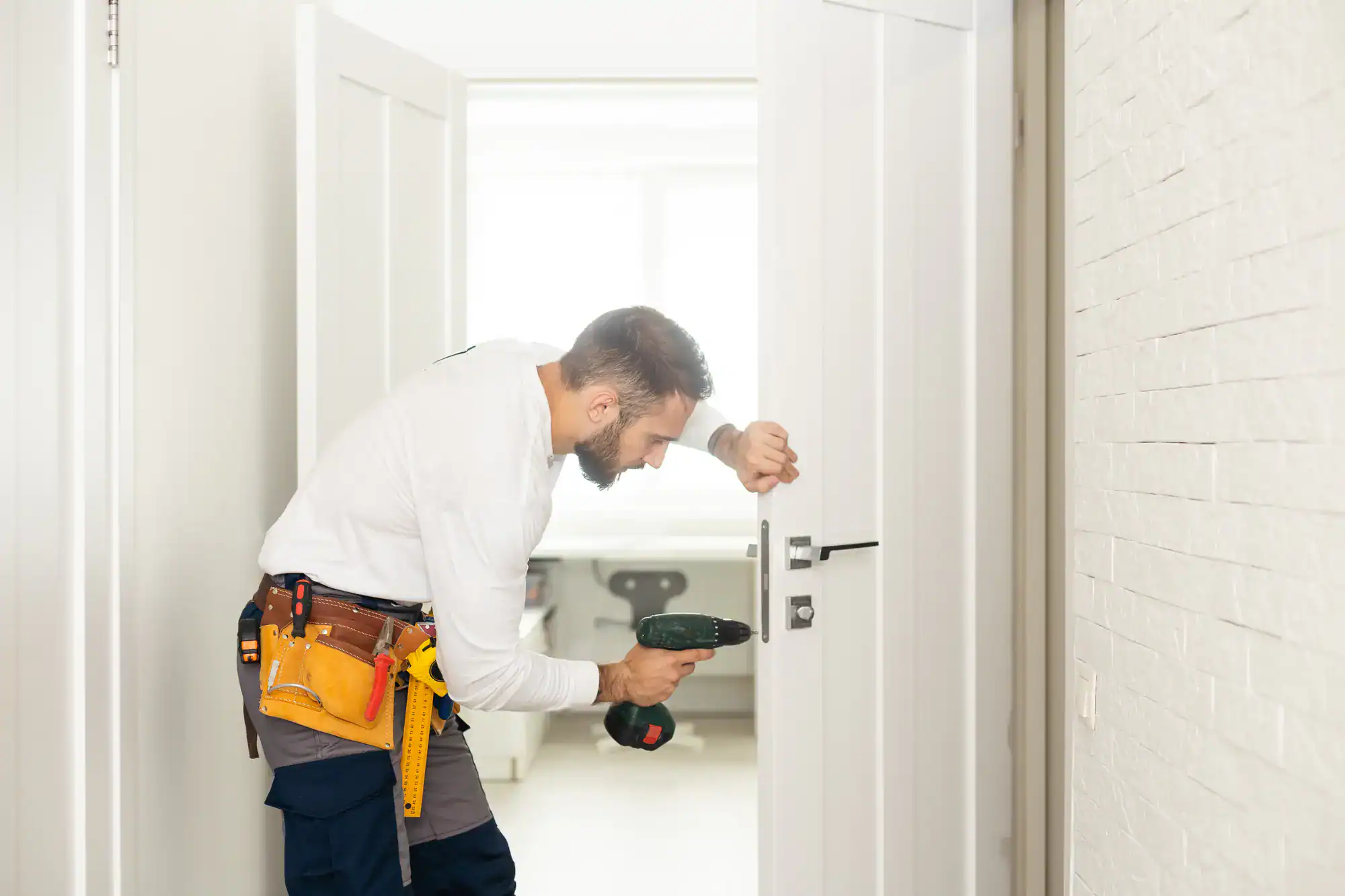 A person in a tool belt uses a power drill to install or repair a door lock in a bright, modern room, showcasing professional locksmith services NYC.