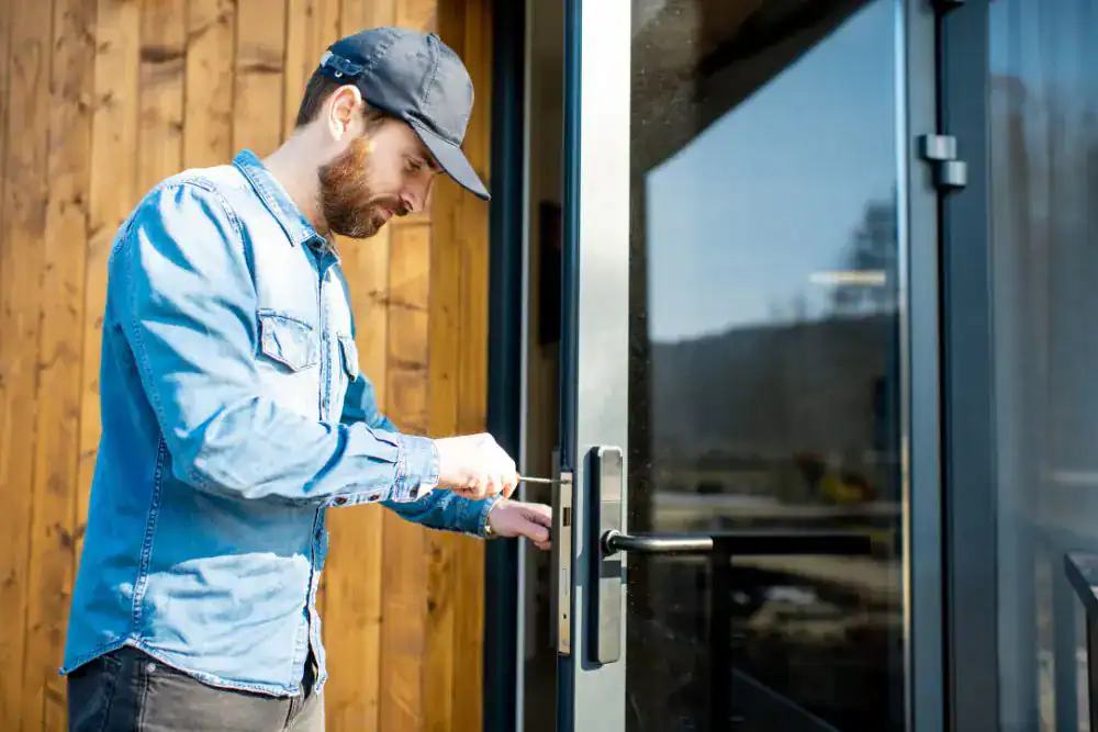 A man in a blue shirt and cap provides locksmith services NYC, using a tool to unlock a glass door outside a modern wooden building in daylight.