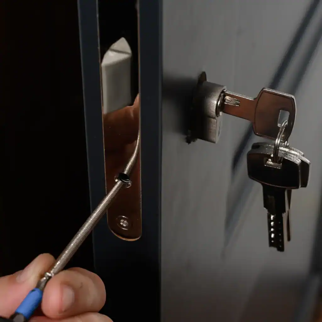 A hand uses a screwdriver to tamper with the lock of a dark-colored door, which has keys inserted into the keyhole on the outside—an urgent situation suited for professional locksmith services NYC.