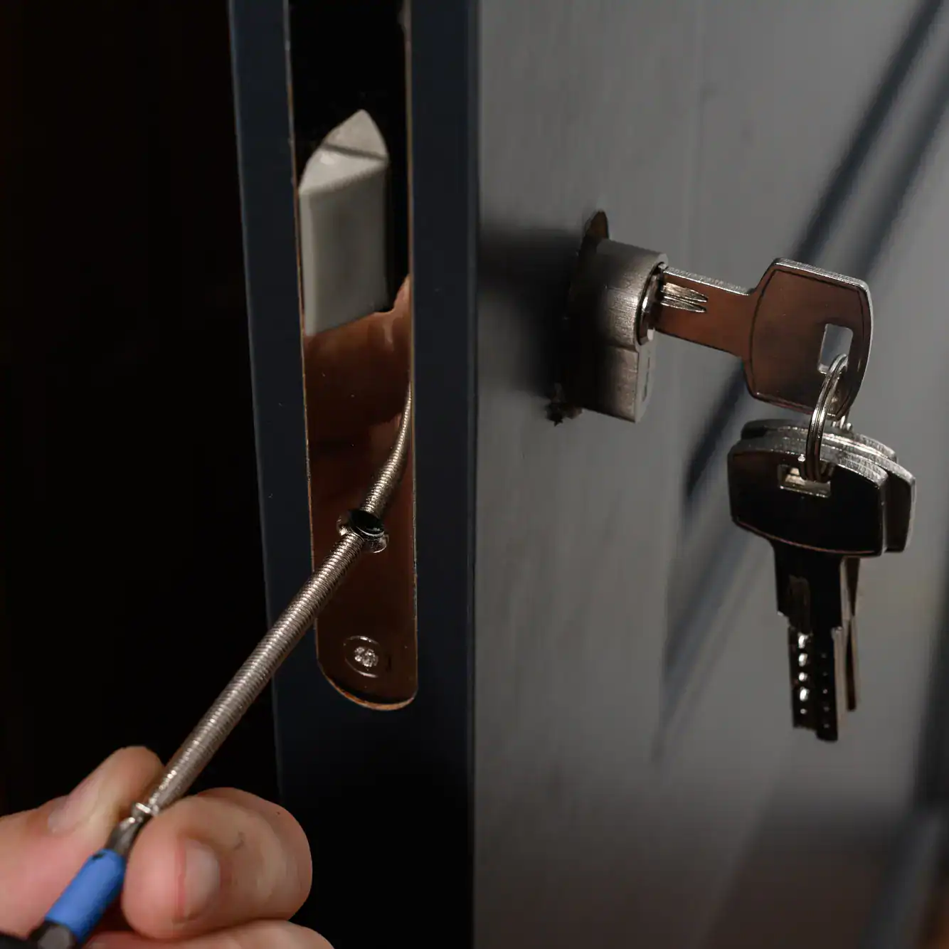A hand uses a screwdriver to tamper with the lock of a dark-colored door, which has keys inserted into the keyhole on the outside—an urgent situation suited for professional locksmith services NYC.